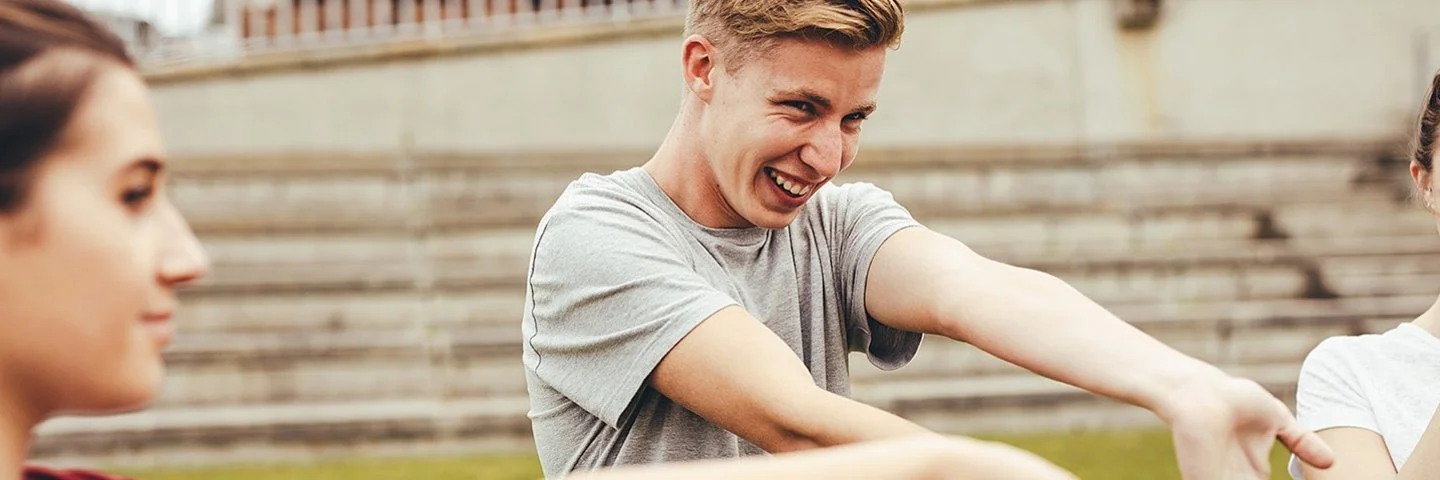 Vor dem Schulgebäude stehen zwei Schülerinnen und ein Schüler im Teenageralter und dehnen ihre Arme.
