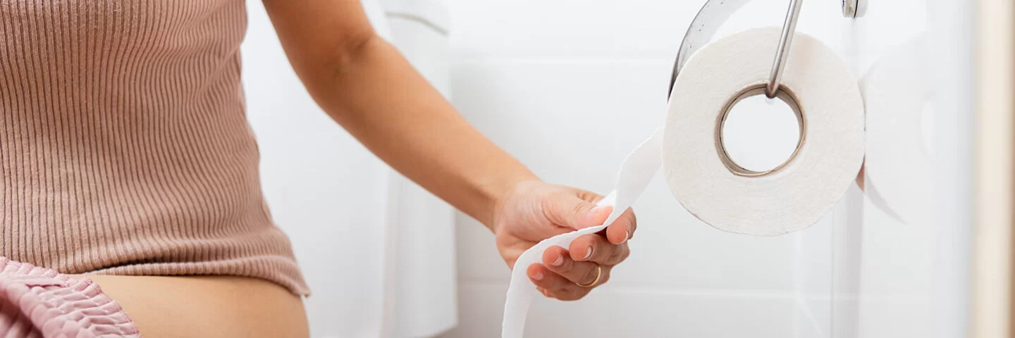 Eine Frau sitzt auf der Toilette und reißt sich ein Stück Toilettenpapier ab.