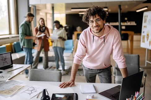 Junger lässig gekleideter Mann steht an seinem Arbeitstisch und schaut in die Kamera.