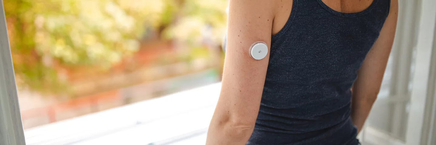 Eine Frau mit einem Sensor für die Blutzuckermessung am Arm schaut aus dem Fenster. Real-Time-Messgeräte sind Hilfsmittel, die die AOK fördert.
