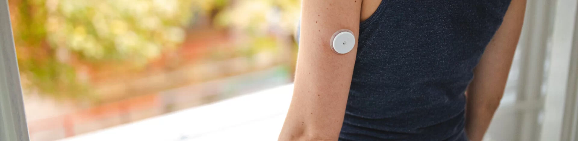 Eine Frau mit einem Sensor für die Blutzuckermessung am Arm schaut aus dem Fenster. Real-Time-Messgeräte sind Hilfsmittel, die die AOK fördert.