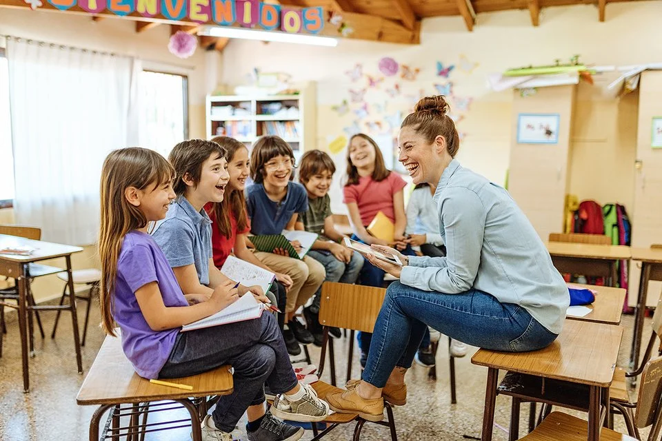 n einem freundlich gestalteten Klassenzimmer sitzt eine Lehrerin im Stuhlkreis mit etwa acht Grundschulkindern. Alle lachen gemeinsam. Die Kinder halten Hefte oder Schreibmaterialien in der Hand. Im Hintergrund sind bunte Wanddekorationen und Regale mit Unterrichtsmaterialien zu sehen.