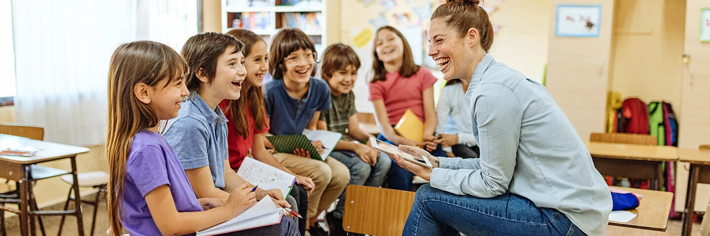 n einem freundlich gestalteten Klassenzimmer sitzt eine Lehrerin im Stuhlkreis mit etwa acht Grundschulkindern. Alle lachen gemeinsam. Die Kinder halten Hefte oder Schreibmaterialien in der Hand. Im Hintergrund sind bunte Wanddekorationen und Regale mit Unterrichtsmaterialien zu sehen.