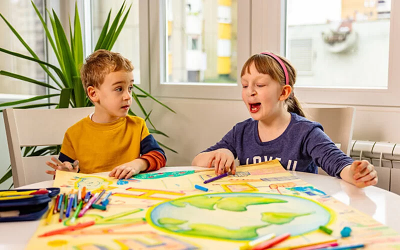 Ein Junge schaut ein Mädchen an, das mit einer Lippen-Kiefer-Gaumen-Spalte (LKGS) neben ihm sitzt und spielt.