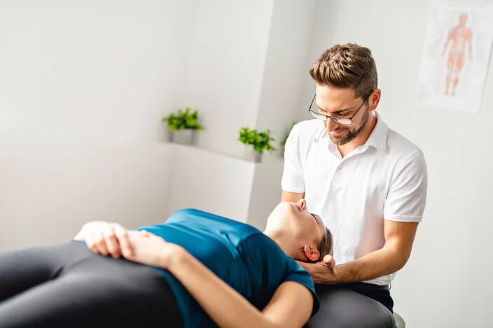 Eine Frau liegt entspannt auf dem Rücken, ein Osteopath sitzt hinter ihrem Kopf und hält seine Hände unter ihren Nacken.