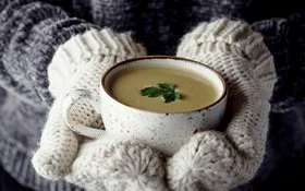 Hände in Wollhandschuhen halten eine dampfende Suppe in schöner Tasse.