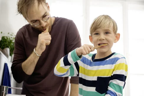 Ein Mann und ein Junge stehen nebenbeinander und halten sich jeweils eine Zahnbürste in den Mund. Das Mundhygienezentrum klärt über das Zähneputzen auf.