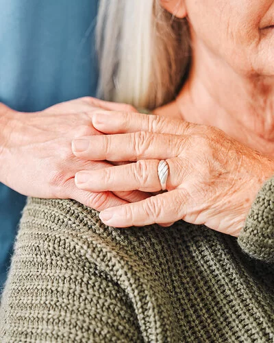 Die Hand einer Pflegerin ruht auf der Schulter einer pflegebedürftigen Frau im Rollstuhl, die ihre Hand auf die Hand der Pflegerin legt.