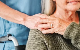 Die Hand einer Pflegerin ruht auf der Schulter einer pflegebedürftigen Frau im Rollstuhl, die ihre Hand auf die Hand der Pflegerin legt.