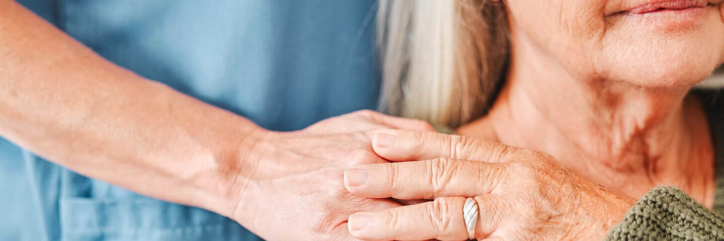 Die Hand einer Pflegerin ruht auf der Schulter einer pflegebedürftigen Frau im Rollstuhl, die ihre Hand auf die Hand der Pflegerin legt.