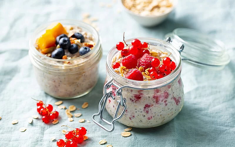 Zwei offene Gläser mit Overnight Oats, gefüllt mit frischen Beeren und weiteren Früchten, auf einer Leinentischdecke. Im Hintergrund eine Schüssel Haferflocken und neben den Gläsern einige rote Johannisbeeren und einzelne Haferflocken.
