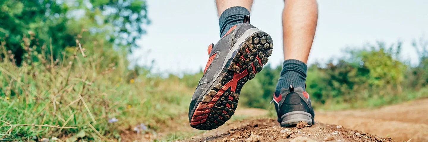 Großaufnahme von Füßen einer Person, die in Trekkingschuhen einen Feldweg entlangläuft. Der linke Fuß ist angehoben und hängt etwas nach unten und zur Seite herab.