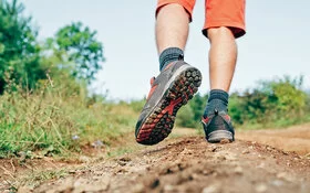 Großaufnahme von Füßen einer Person, die in Trekkingschuhen einen Feldweg entlangläuft. Der linke Fuß ist angehoben und hängt etwas nach unten und zur Seite herab.
