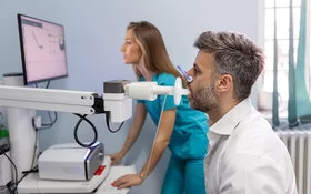 Ein Mann beim Lungenfunktionstest mit einem Spirometer, im Hintergrund eine medizinische Fachangestellte.