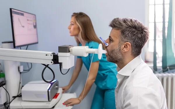 Ein Mann beim Lungenfunktionstest mit einem Spirometer, im Hintergrund eine medizinische Fachangestellte.