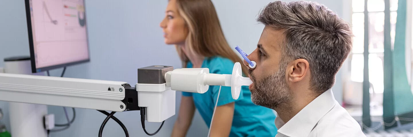Ein Mann beim Lungenfunktionstest mit einem Spirometer, im Hintergrund eine medizinische Fachangestellte.