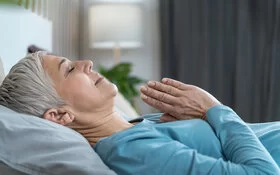 Eine grauhaarige ältere Frau liegt mit geschlossenen Augen und mit vor der Brust gefalteten Händen im Bett und meditiert vor dem Einschlafen.
