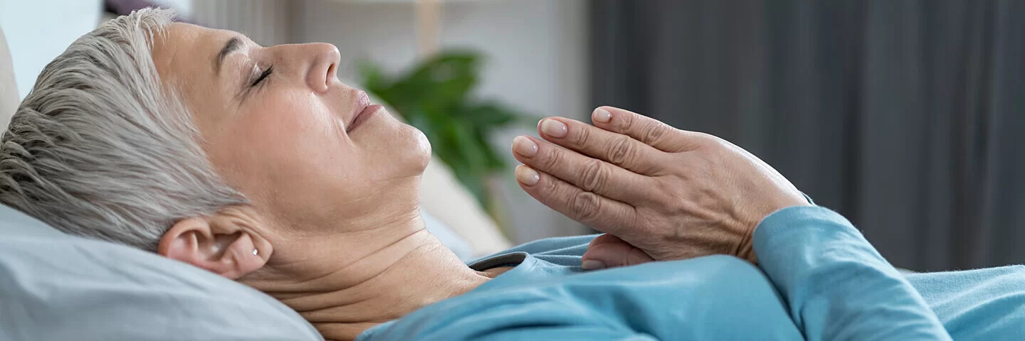 Eine grauhaarige ältere Frau liegt mit geschlossenen Augen und mit vor der Brust gefalteten Händen im Bett und meditiert vor dem Einschlafen.