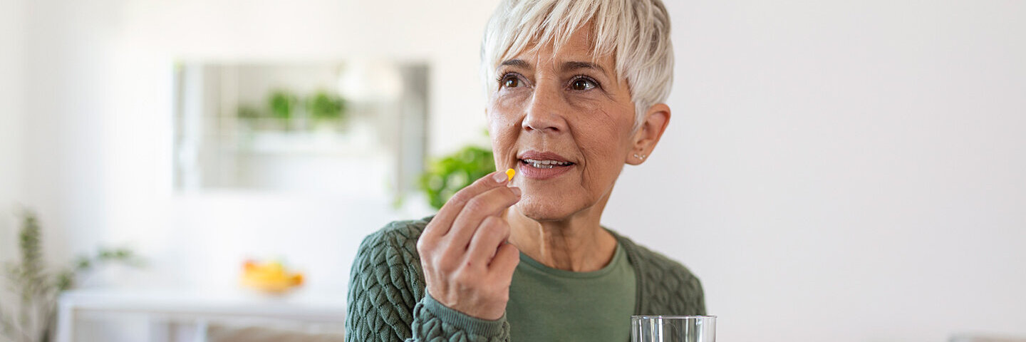 Ältere Frau hält ein Glas Wasser in der Hand und führt eine Tablette zum Mund.