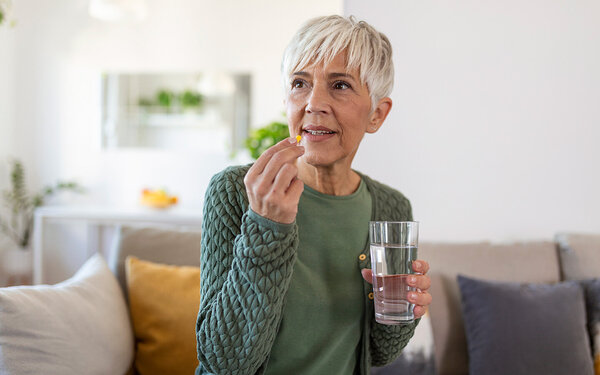 Ältere Frau hält ein Glas Wasser in der Hand und führt eine Tablette zum Mund.