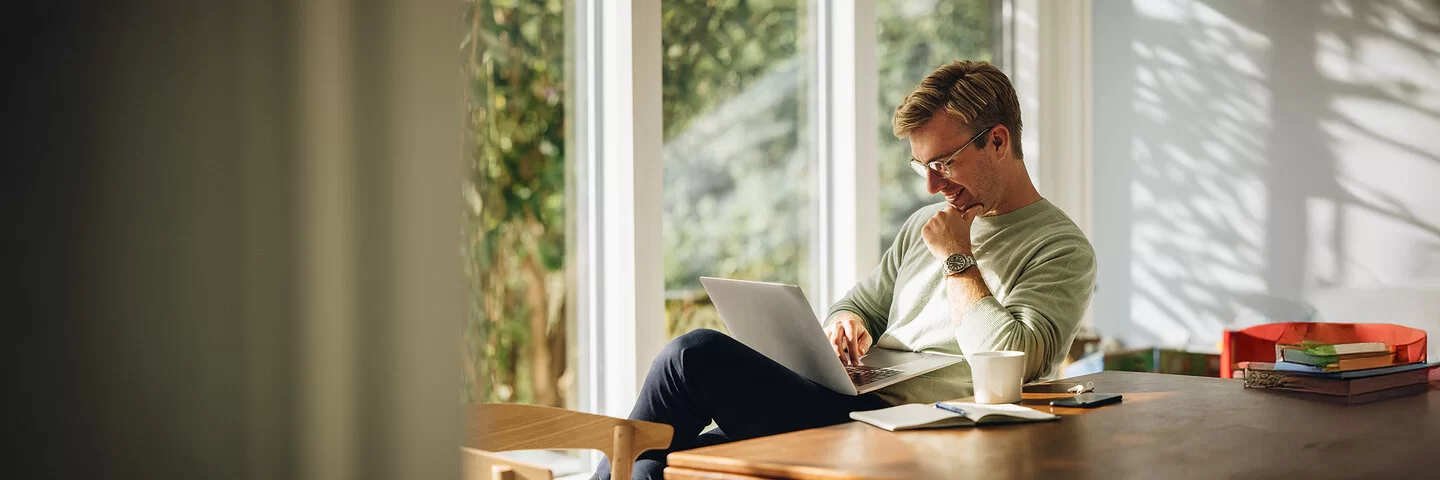 Ein Mann mittleren Alters mit Brille, grauem Sweatshirt und dunkler Hose sitzt mit überschlagenen Beinen an einem Tisch vor dem Fenster. Sein Laptop liegt auf seinem Schoß.