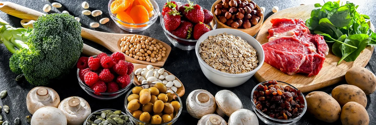 Auf einem Tisch sind verschiedene frische Lebensmittel wie Brokkoli, Beeren, Nüsse, Haferflocken und Fleisch arrangiert.