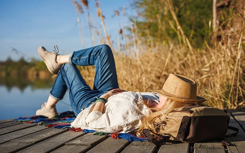 An einem sonnigen Tag liegt eine Frau auf einem Steg an einem See. Sie hat einen Strohhut über das Gesicht gezogen und schlägt ein Bein über das andere.