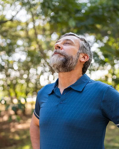 Ein mittelalter Mann steht im Wald und atmet mit geschlossenen Augen tief ein – gezieltes Training und Lungensport stärken die Lunge.