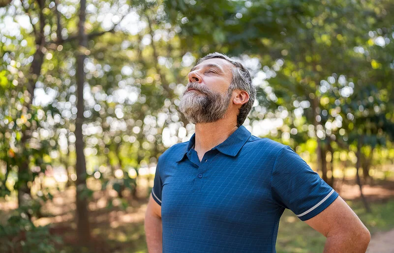 Ein mittelalter Mann steht im Wald und atmet mit geschlossenen Augen tief ein – gezieltes Training und Lungensport stärken die Lunge.