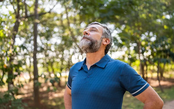Ein mittelalter Mann steht im Wald und atmet mit geschlossenen Augen tief ein – gezieltes Training und Lungensport stärken die Lunge.