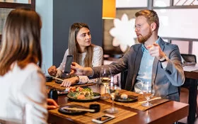 Ein Mann erklärt mit erhobenem Zeigefinger zwei Frauen am Tisch etwas und zeigt dabei als Beweis sein Smartphone herum.