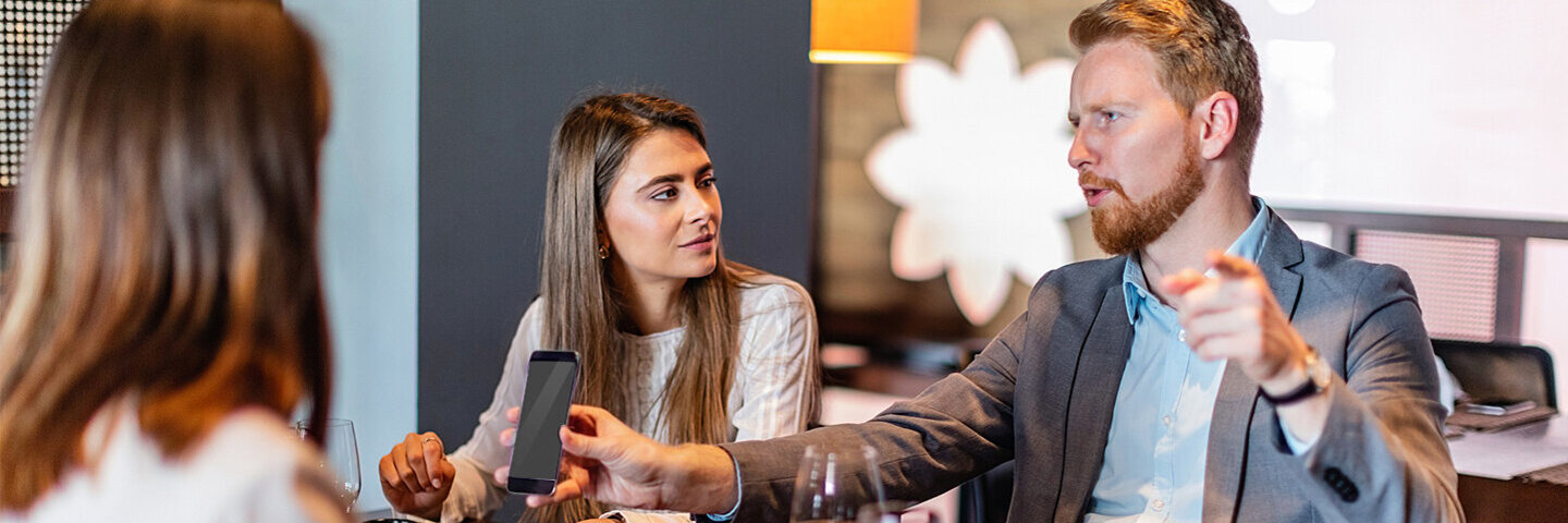 Ein Mann erklärt mit erhobenem Zeigefinger zwei Frauen am Tisch etwas und zeigt dabei als Beweis sein Smartphone herum.