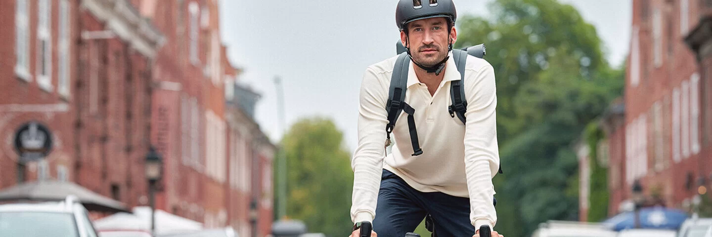 Ein sportlicher junger Mann fährt mit dem Fahrrad durch eine Wohnstraße, rechts und links am Straßenrand parken Autos. Er trägt ein weißes langärmliges Polo-Shirt, eine dunkelblaue Hose, weiße Sneaker und einen schwarzen Fahrradhelm. Er schaut direkt in die Kamera.