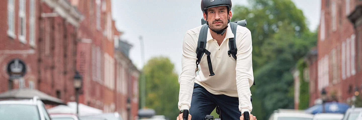 Ein sportlicher junger Mann fährt mit dem Fahrrad durch eine Wohnstraße, rechts und links am Straßenrand parken Autos. Er trägt ein weißes langärmliges Polo-Shirt, eine dunkelblaue Hose, weiße Sneaker und einen schwarzen Fahrradhelm. Er schaut direkt in die Kamera.