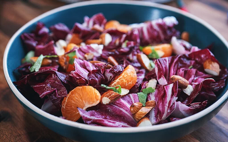 Auf einem Holzbrett steht eine kleine Schale. Darin sind Radicchio, Mandarinen, Mandeln und Salatblätter.