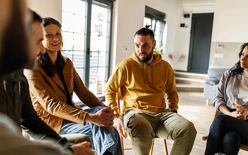 Zwei Männer und zwei Frauen sitzen zusammen in einem Stuhlkreis in einem Beratungsgespräch oder einer Gruppentherapie.