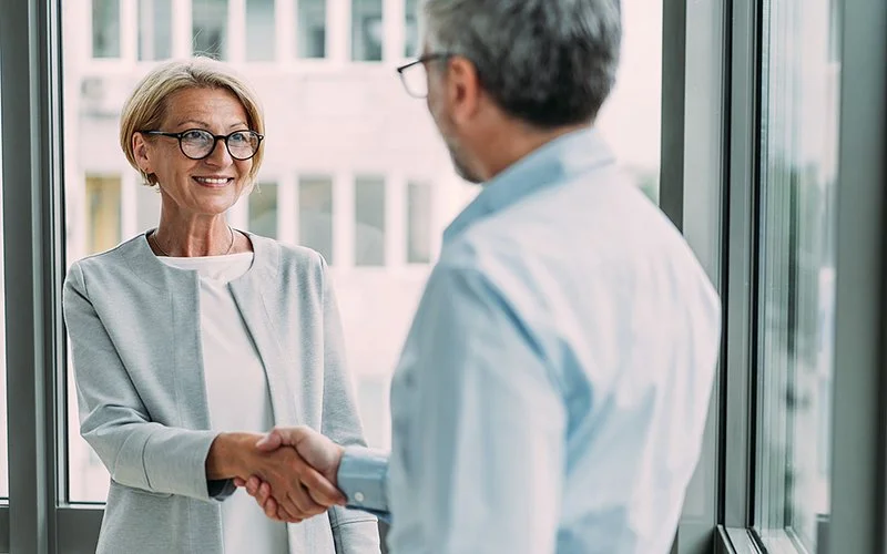 Zwei Personen im Business-Outfit geben sich in einem modernen Büro mit großen Fenstern die Hand.