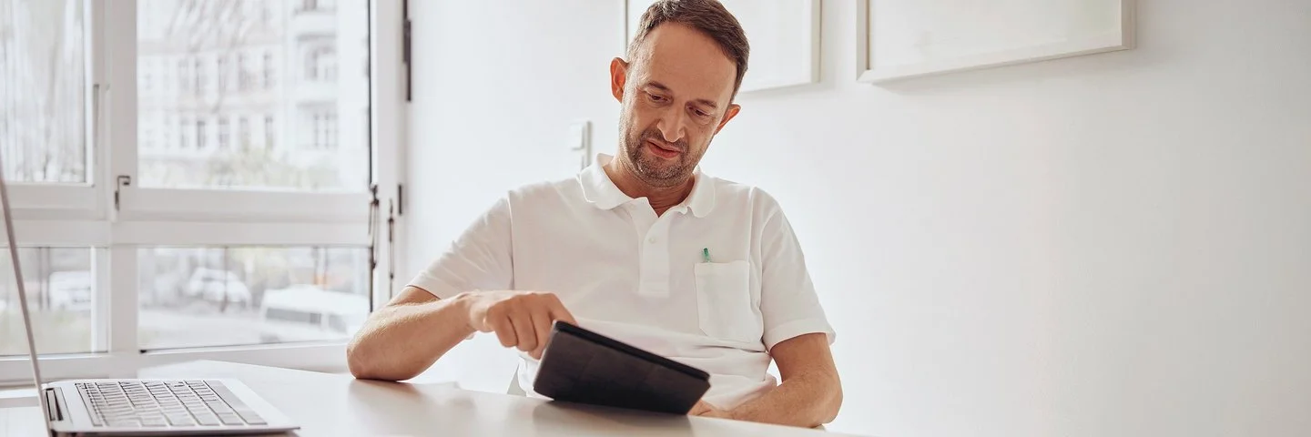 Ein Arzt sitzt an seinem Schreibtisch und schaut auf ein Tablet, neben sich das geöffnete Laptop.