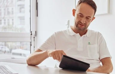 Ein Arzt sitzt an seinem Schreibtisch und schaut auf ein Tablet, neben sich das geöffnete Laptop.