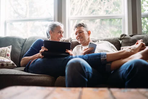 Ein Pärchen in seinen 50ern lümmelt gemeinsam auf der Couch – sie hält ein Tablet, er ein Smartphone in Händen.