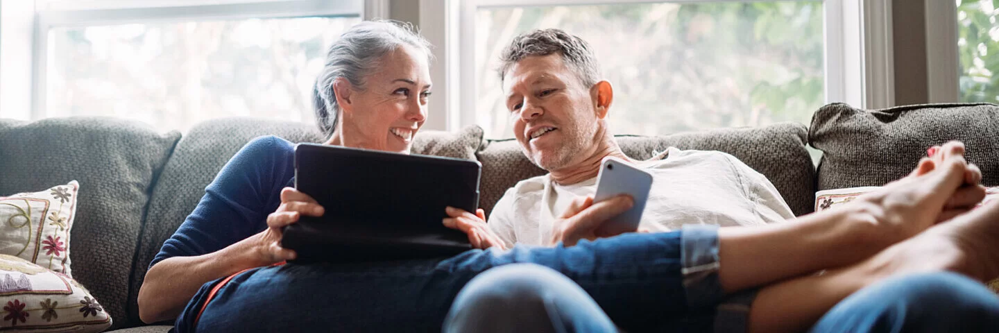 Ein Pärchen in seinen 50ern lümmelt gemeinsam auf der Couch – sie hält ein Tablet, er ein Smartphone in Händen.