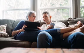 Ein Pärchen in seinen 50ern lümmelt gemeinsam auf der Couch – sie hält ein Tablet, er ein Smartphone in Händen.