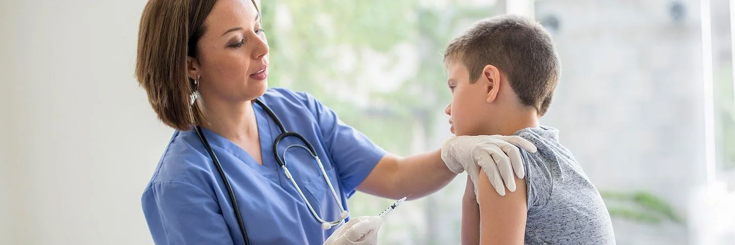 Eine Ärztin oder eine andere weibliche medizinische Fachkraft steht vor einem Jungen, der auf einer Behandlungsbank sitzt. Die Ärztin hält in der rechten Hand eine Spritze und drückt mit den Fingern der linken Hand die Haut im oberen Bereich seines Oberarms zusammen.
