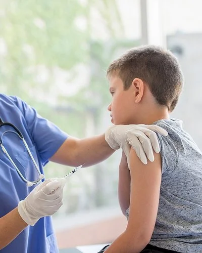 Eine Ärztin oder eine andere weibliche medizinische Fachkraft steht vor einem Jungen, der auf einer Behandlungsbank sitzt. Die Ärztin hält in der rechten Hand eine Spritze und drückt mit den Fingern der linken Hand die Haut im oberen Bereich seines Oberarms zusammen.