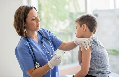 Eine Ärztin oder eine andere weibliche medizinische Fachkraft steht vor einem Jungen, der auf einer Behandlungsbank sitzt. Die Ärztin hält in der rechten Hand eine Spritze und drückt mit den Fingern der linken Hand die Haut im oberen Bereich seines Oberarms zusammen.