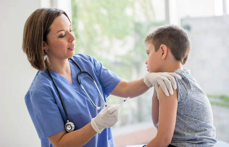 Eine Ärztin oder eine andere weibliche medizinische Fachkraft steht vor einem Jungen, der auf einer Behandlungsbank sitzt. Die Ärztin hält in der rechten Hand eine Spritze und drückt mit den Fingern der linken Hand die Haut im oberen Bereich seines Oberarms zusammen.