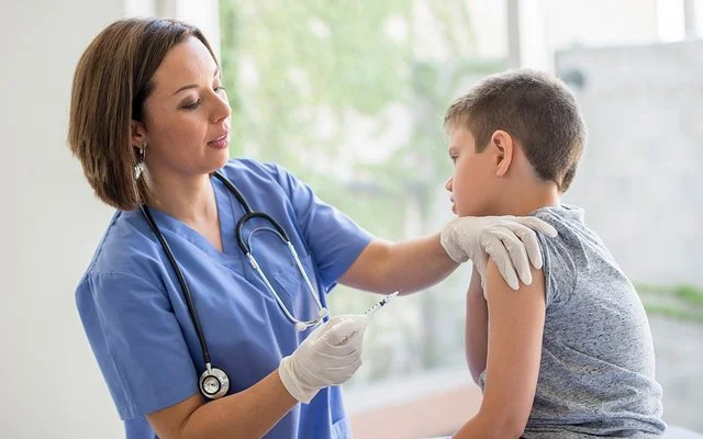 Eine Ärztin oder eine andere weibliche medizinische Fachkraft steht vor einem Jungen, der auf einer Behandlungsbank sitzt. Die Ärztin hält in der rechten Hand eine Spritze und drückt mit den Fingern der linken Hand die Haut im oberen Bereich seines Oberarms zusammen.