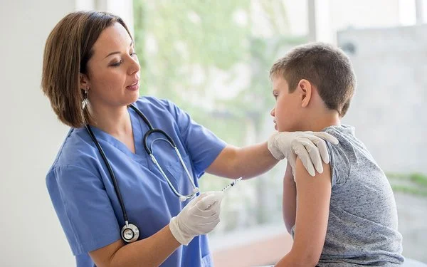 Eine Ärztin oder eine andere weibliche medizinische Fachkraft steht vor einem Jungen, der auf einer Behandlungsbank sitzt. Die Ärztin hält in der rechten Hand eine Spritze und drückt mit den Fingern der linken Hand die Haut im oberen Bereich seines Oberarms zusammen.