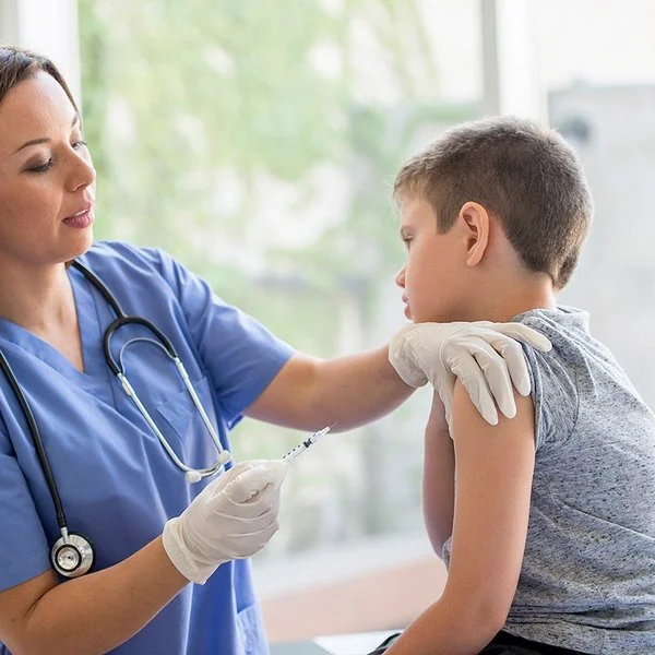 Eine Ärztin oder eine andere weibliche medizinische Fachkraft steht vor einem Jungen, der auf einer Behandlungsbank sitzt. Die Ärztin hält in der rechten Hand eine Spritze und drückt mit den Fingern der linken Hand die Haut im oberen Bereich seines Oberarms zusammen.