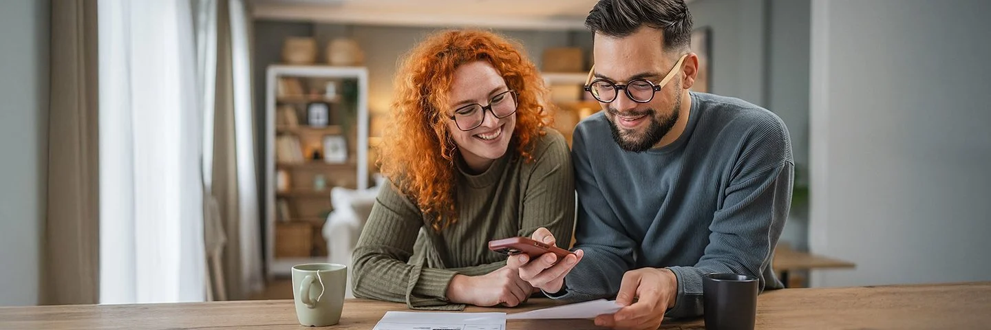 Zwei Personen sitzen nebeneinander an einem Holztisch. Beide beugen sich leicht nach vorne und halten jeweils ein Blatt Papier in der Hand. Eine Person zeigt mit einem Smartphone auf das Dokument. Im Hintergrund sind Regale mit Dekorationsgegenständen und ein Fenster mit weißen Vorhängen zu sehen.